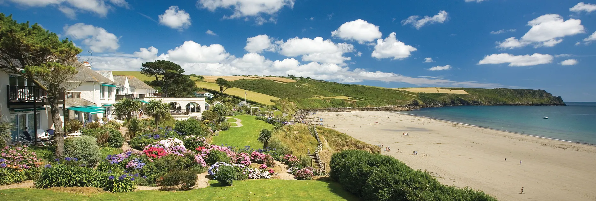 Coastal hotel and colourful gardens overlooking a sandy beach and headland under a bright blue sky