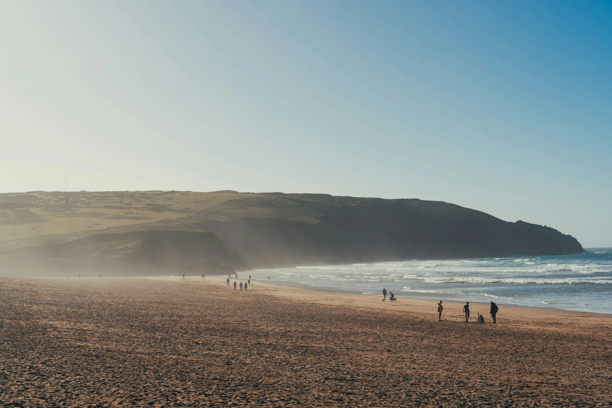 Wide sandy beach with scattered people walking along the shoreline beneath cliffs and a clear blue sky