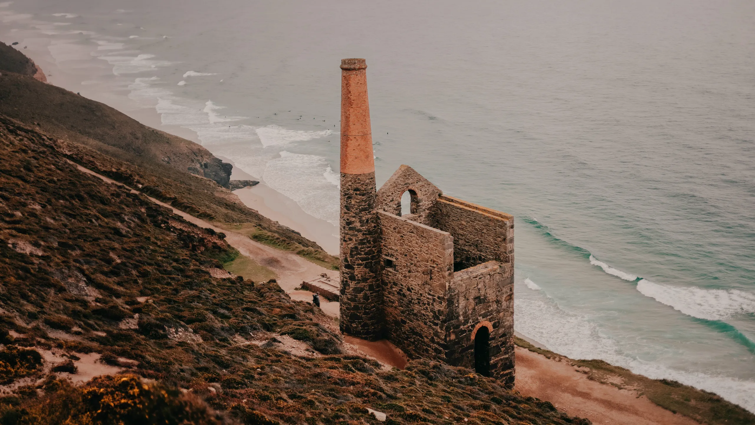 Ruined engine house with a tall chimney on a clifftop above the sea and sandy beach.