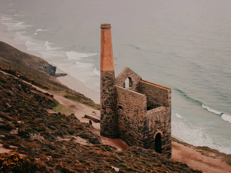 Ruined engine house with a tall chimney on a clifftop above the sea and sandy beach.