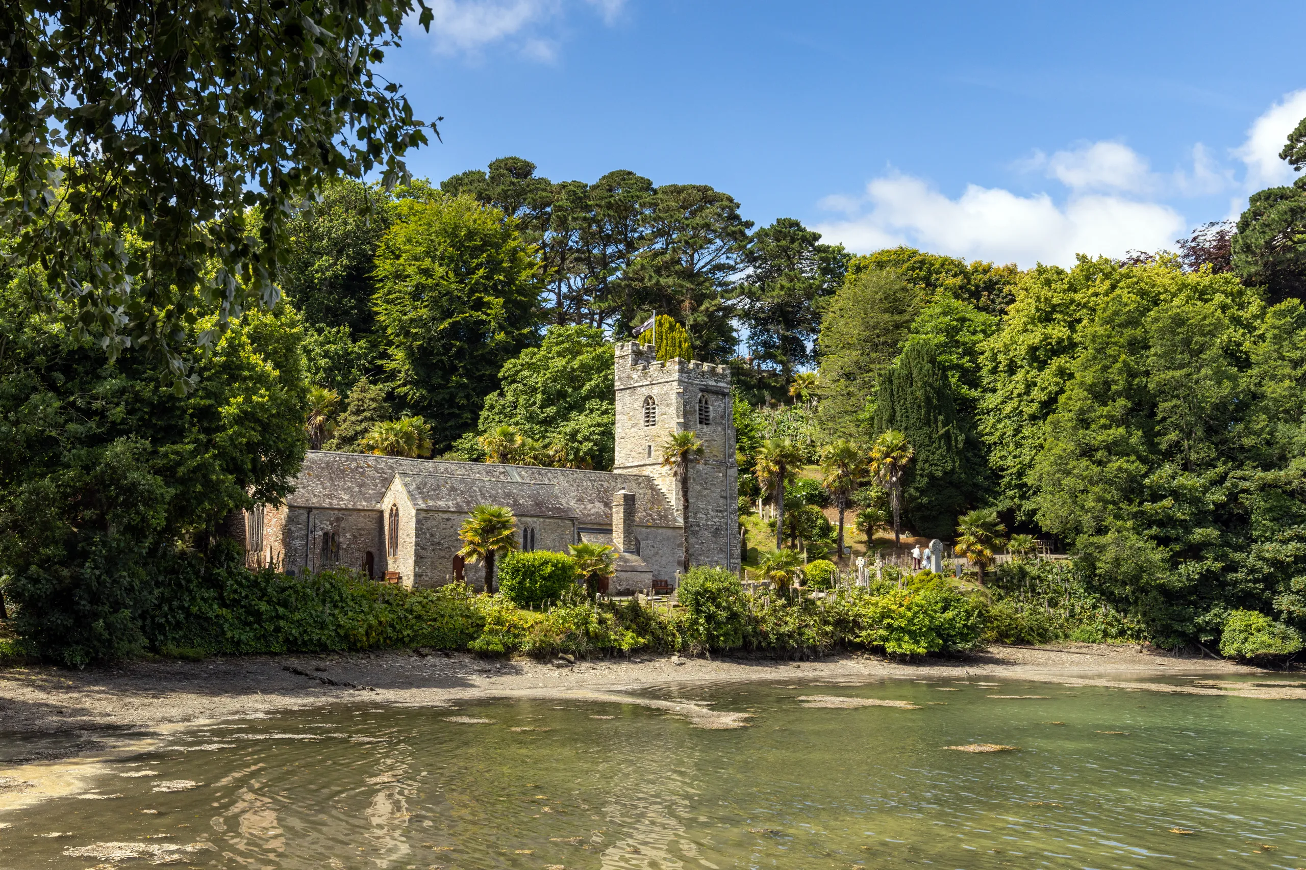 Stone church by a sheltered waterfront surrounded by dense green trees under a blue sky