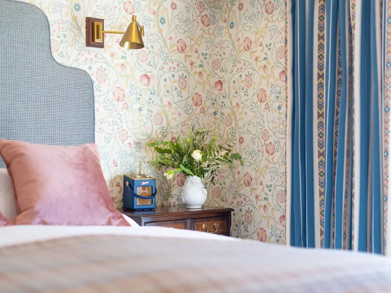 Stylish bedroom corner with a blue upholstered headboard, pink velvet cushions, floral wallpaper and striped blue curtains.