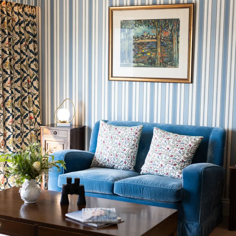 Elegant sitting room with a blue upholstered sofa, floral cushions, striped wallpaper and a wooden coffee table.