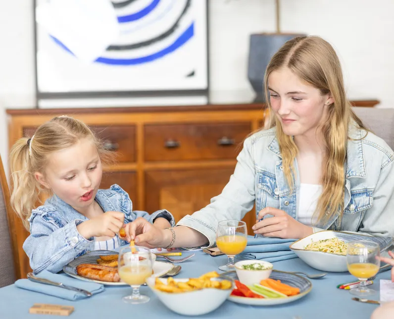 Two girls eating a meal together at a dining table with sausages, chips, vegetables and glasses of orange juice