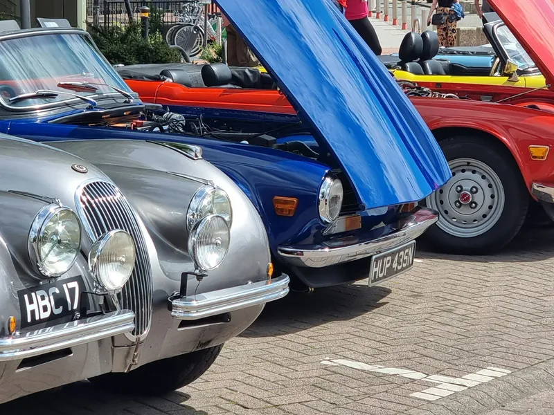 A row of classic cars with raised bonnets parked at an outdoor car show on a sunny day.