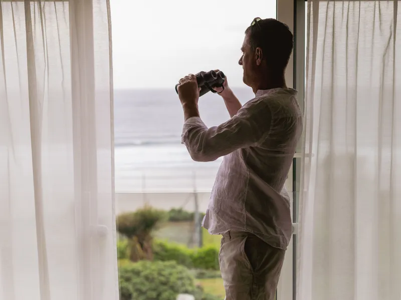 Man standing by a window with binoculars, looking out towards the sea through sheer curtains