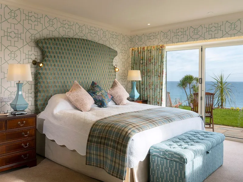 Elegant seaside hotel suite bedroom with a patterned headboard, blue accents and sliding doors opening to an ocean view terrace.