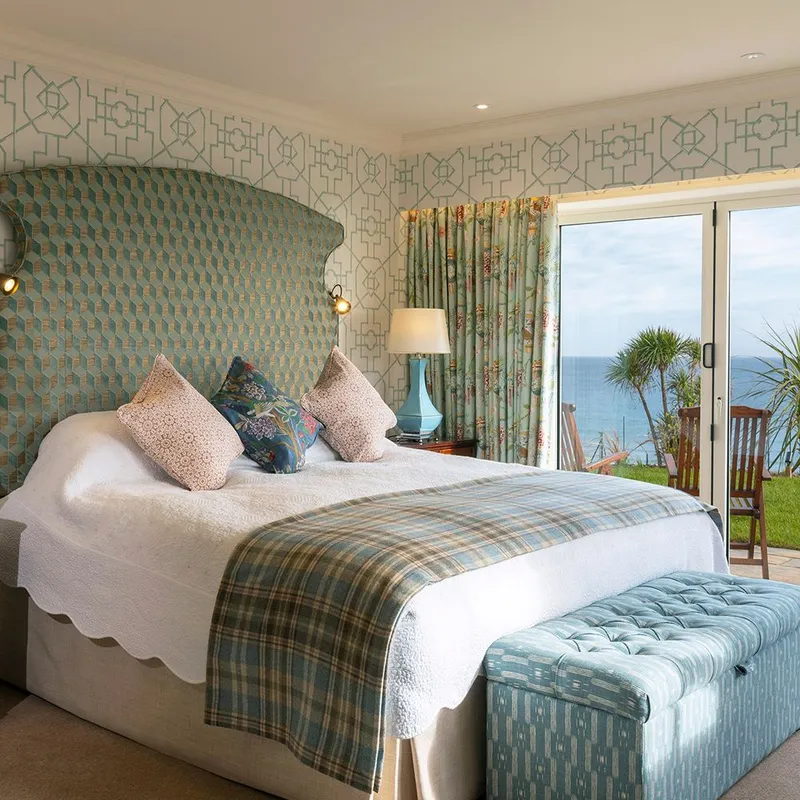 Elegant seaside hotel suite bedroom with a patterned headboard, blue accents and sliding doors opening to an ocean view terrace.