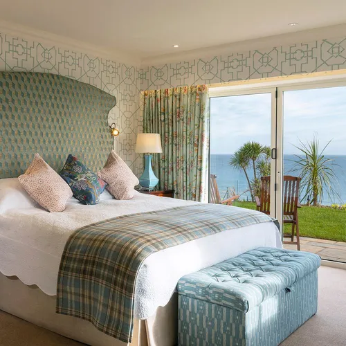 Elegant seaside hotel suite bedroom with a patterned headboard, blue accents and sliding doors opening to an ocean view terrace.