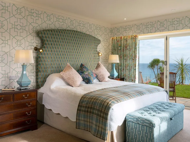 Elegant seaside hotel suite bedroom with a patterned headboard, blue accents and sliding doors opening to an ocean view terrace.