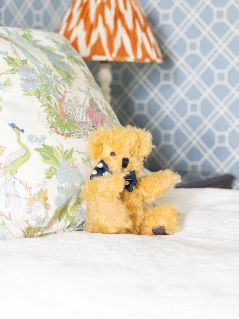 Cosy bedroom with blue geometric wallpaper, floral cushion and a teddy bear on a white bed