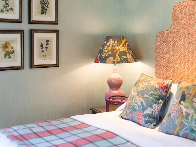 Cosy bedroom corner with a patterned upholstered headboard, floral cushions, a pink ceramic lamp and framed botanical prints.