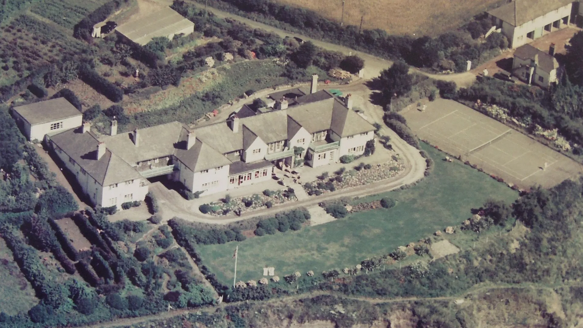 Historic aerial view of The Nare hotel and grounds beside the Cornish coastline, with tennis court and surrounding gardens