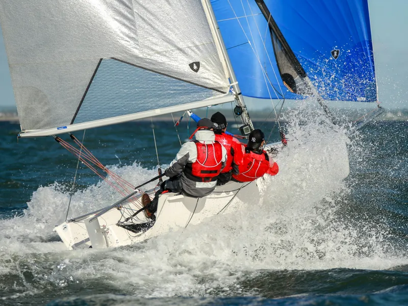 Three sailors in red life jackets racing a small sailboat through choppy water with blue and grey sails