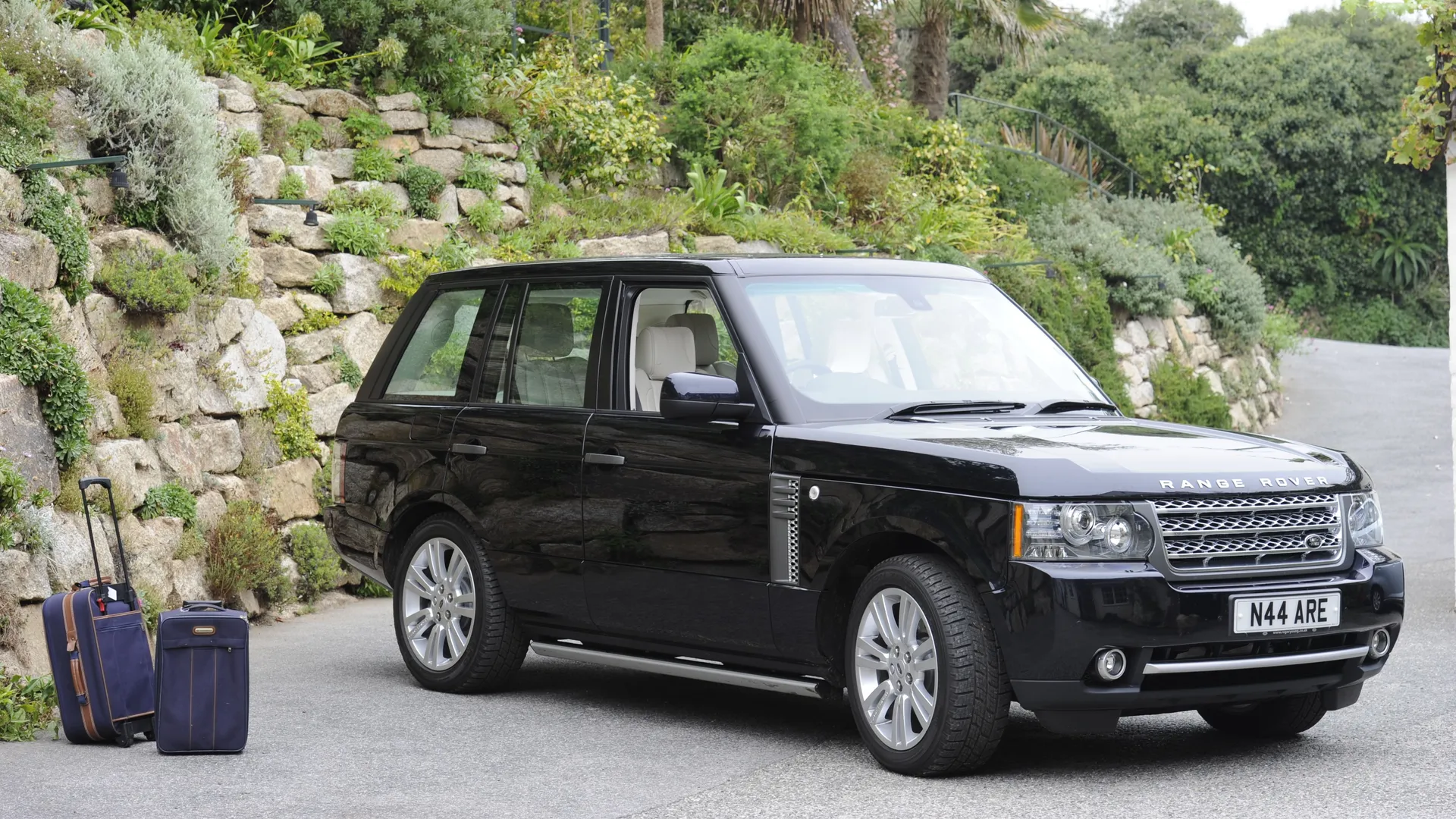 Black Range Rover parked beside a stone garden wall with two suitcases on the roadside