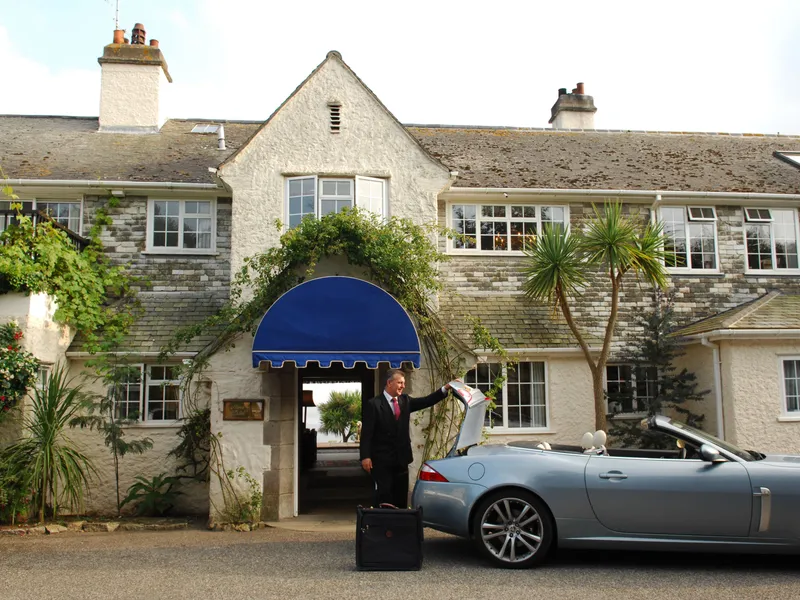 Man in a suit unloading luggage from a silver convertible outside a traditional stone hotel with a blue entrance canopy.