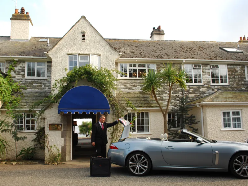 Man in a suit unloading luggage from a silver convertible outside a traditional stone hotel with a blue entrance canopy.