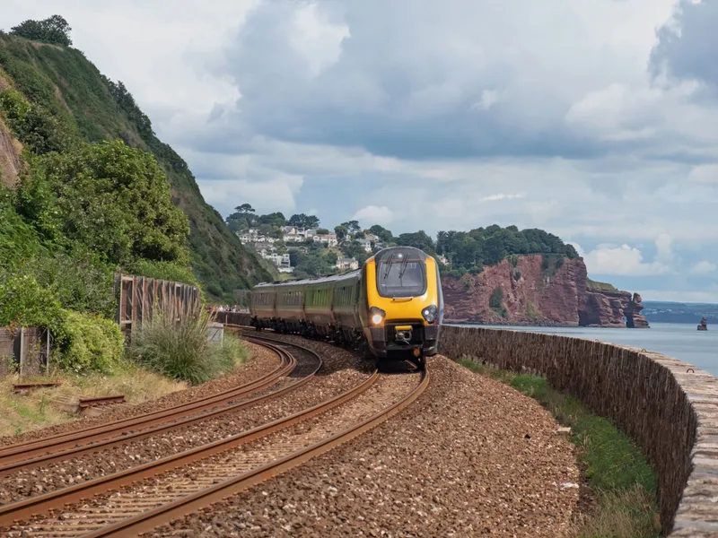 Passenger train travelling along a curved coastal railway beside red cliffs and the sea under dramatic clouds