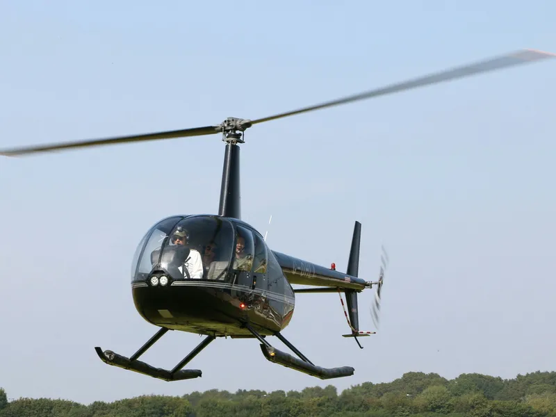 Black helicopter hovering above a grassy airfield under a clear blue sky