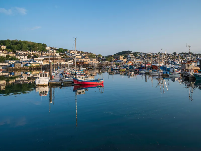 Newlyn Fishing boats and small yachts moored in a calm harbour with reflections on the water and hillside houses under a clear blue sky.