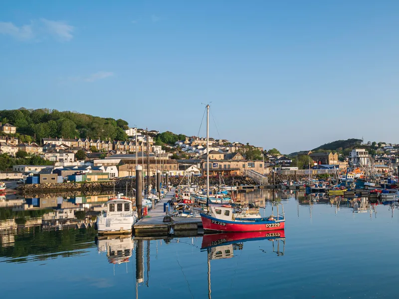 Newlyn Fishing boats and small yachts moored in a calm harbour with reflections on the water and hillside houses under a clear blue sky.