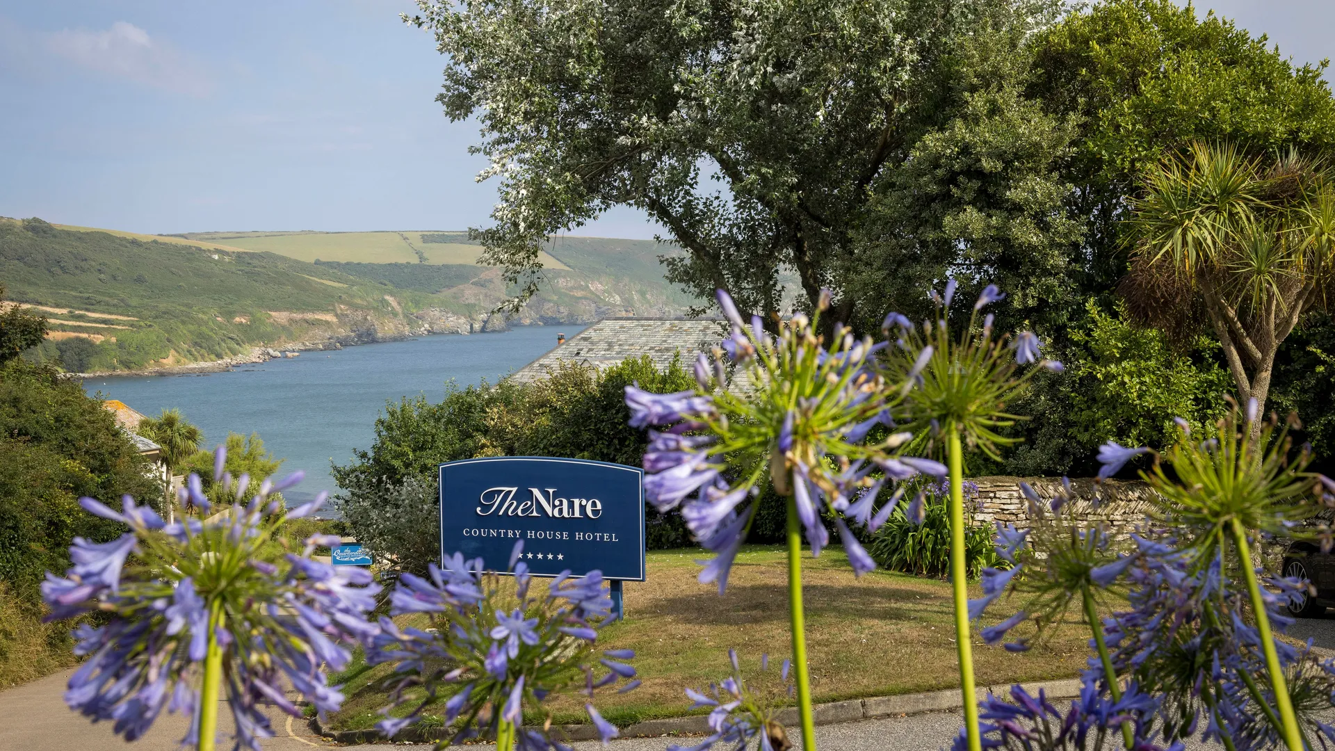The Nare Country House Hotel sign with purple agapanthus flowers in the foreground and a coastal bay view behind