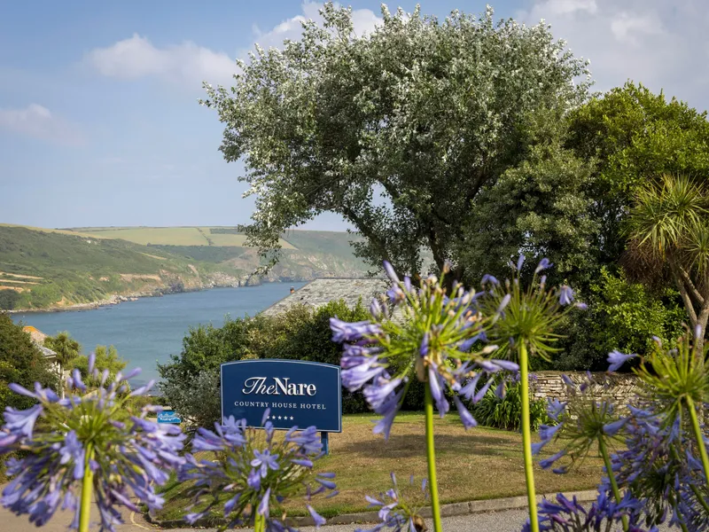 The Nare Country House Hotel sign with purple agapanthus flowers in the foreground and a coastal bay view behind
