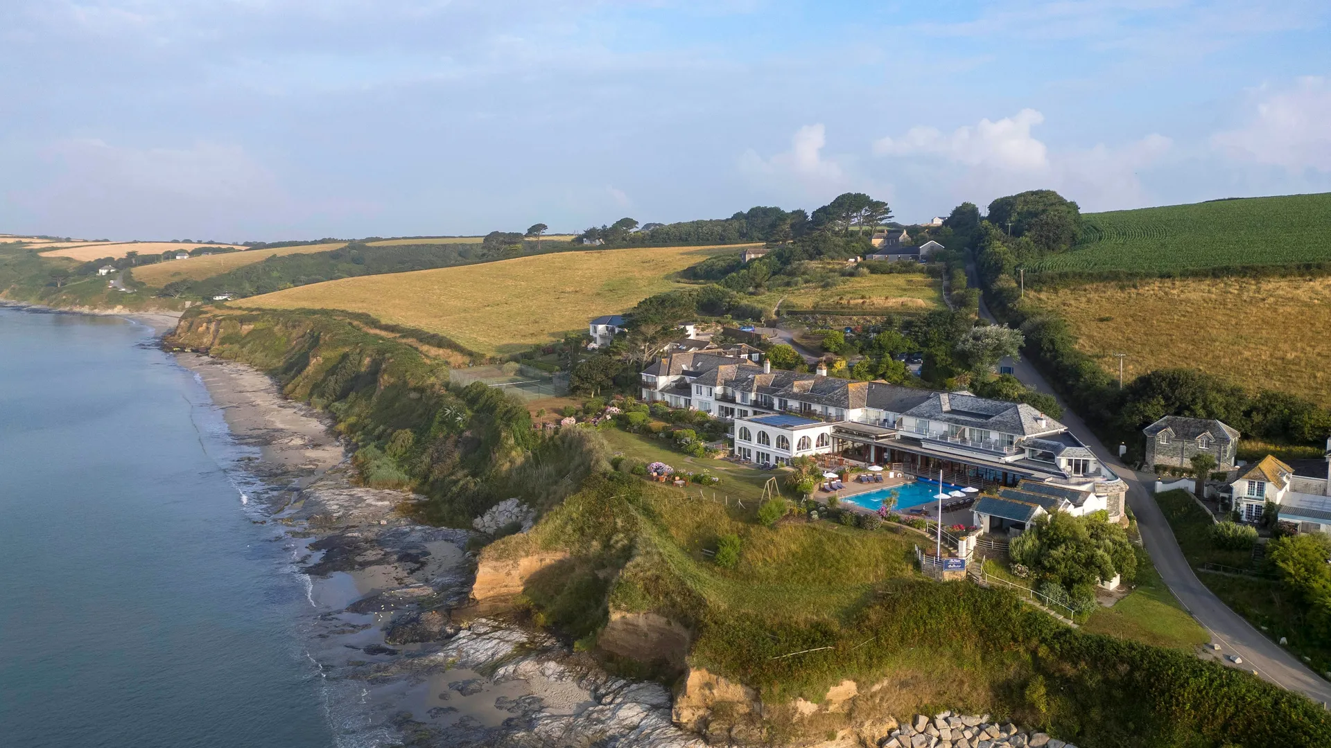 Aerial view of a coastal hotel on clifftops beside a sandy beach and calm sea, surrounded by rolling countryside.