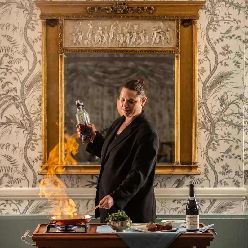 Chef flambéing a dish beside a gilded mirror in an elegant wallpapered dining room