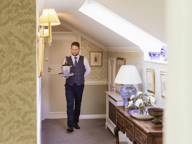 A hotel staff member carrying a tray with champagne and ice bucket along an elegant corridor.