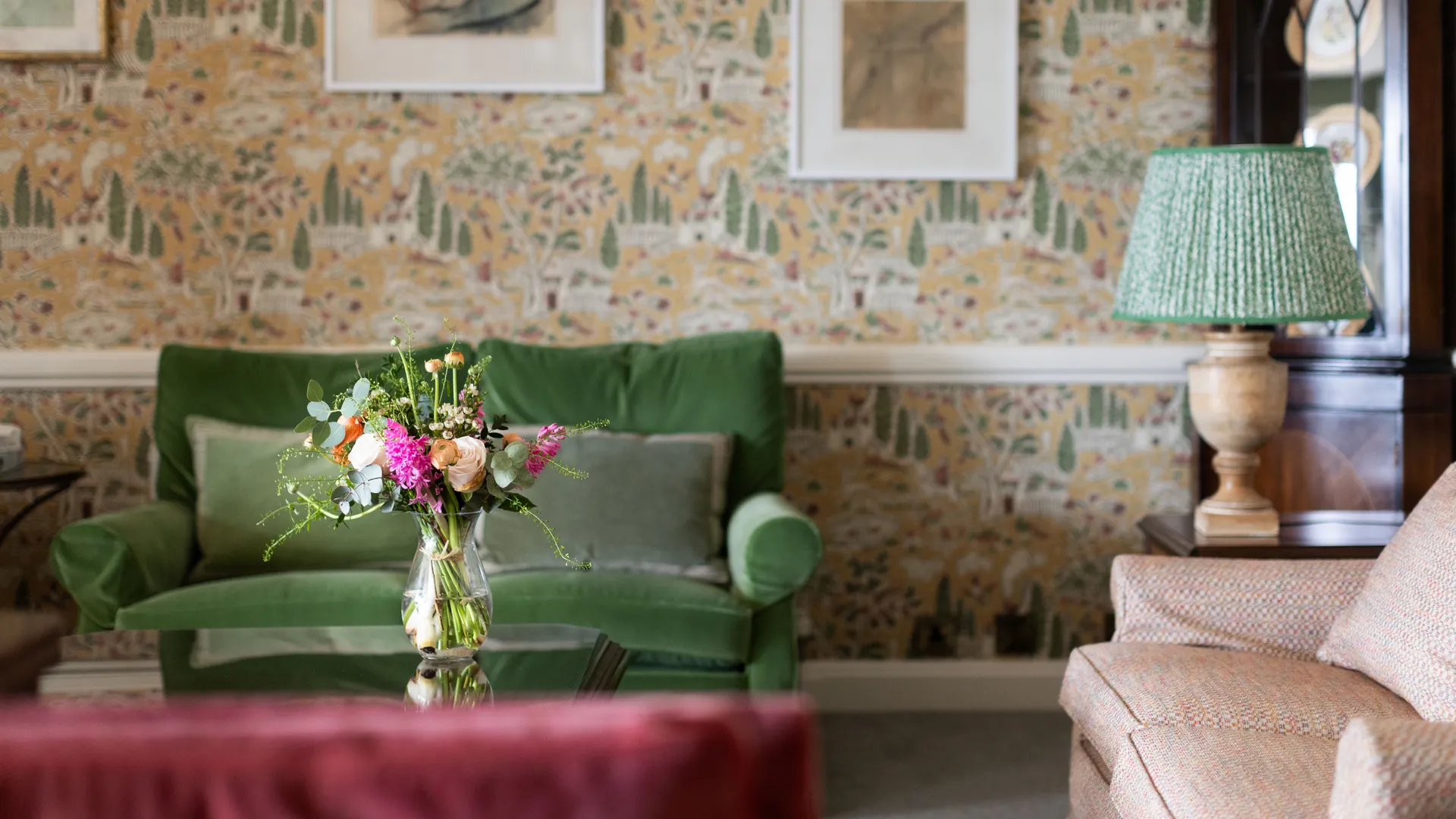 Cosy sitting room with patterned wallpaper, a green sofa, floral arrangement on a glass coffee table, and a table lamp beside a dark wooden cabinet