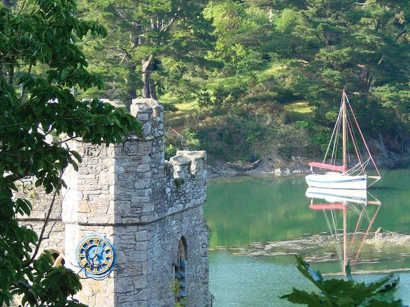 St Just in Roseland Church Stone tower with a clock beside a calm inlet, with a white sailing boat reflected in the water and trees surrounding the scene.