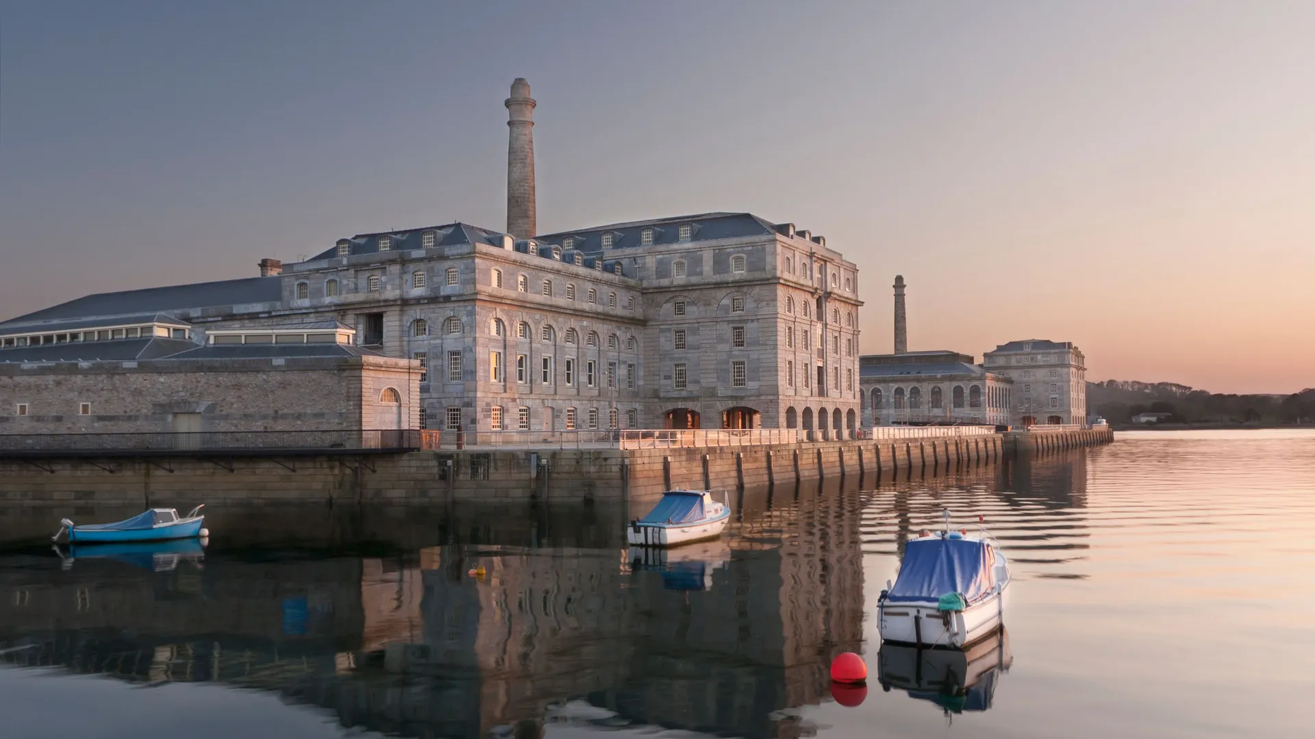 Historic waterfront building with tall chimney reflected in calm harbour water, with small boats moored at sunset.
