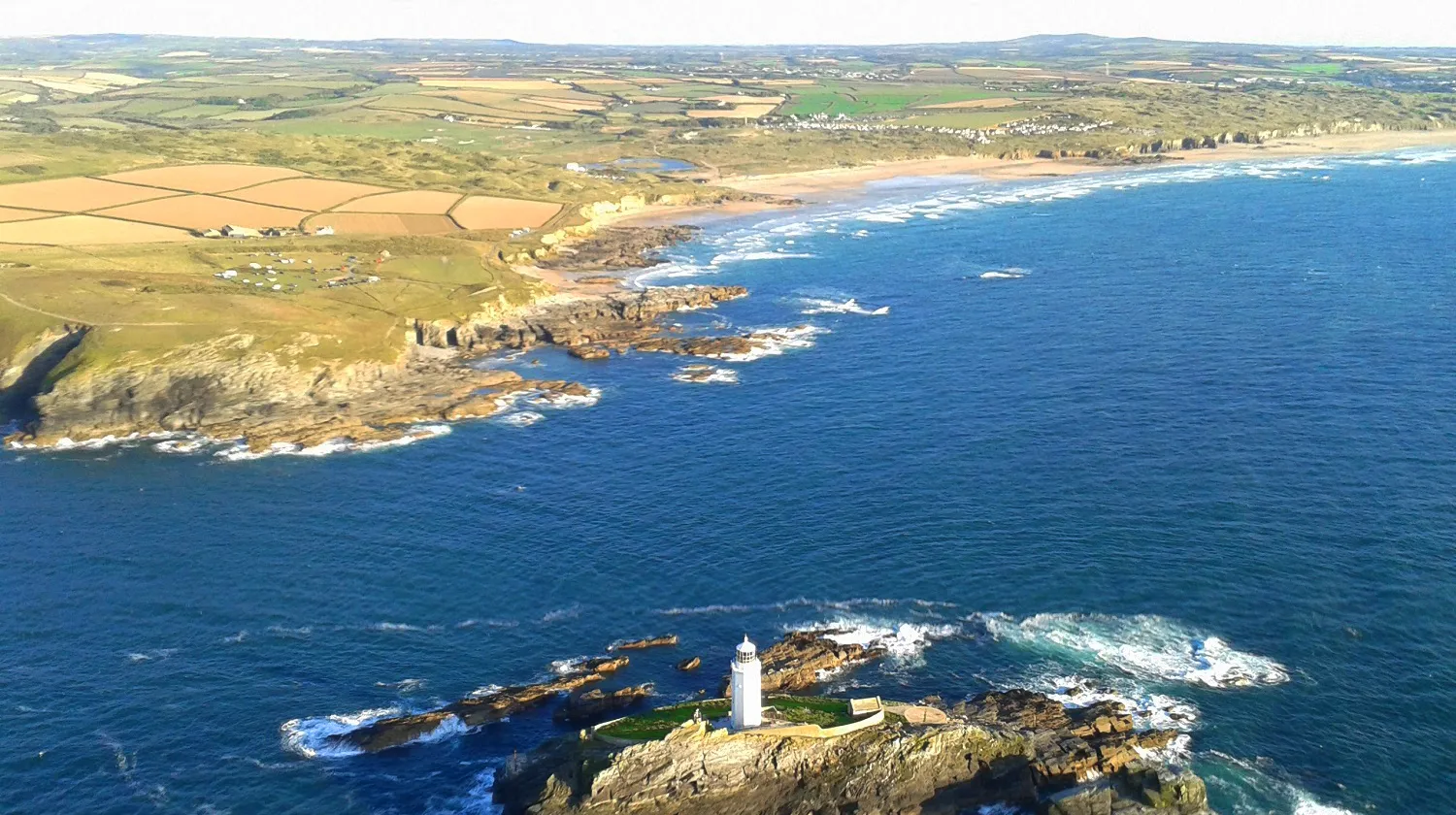 Aerial view of Godrevy Lighthouse on a rocky island off the Cornish coast, with blue sea, sandy bays and patchwork fields inland.