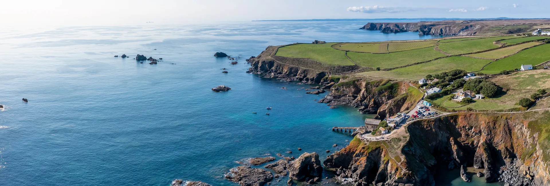Aerial view of a rugged coastal bay with turquoise water, rocky cliffs, scattered houses and green fields stretching along the shoreline.