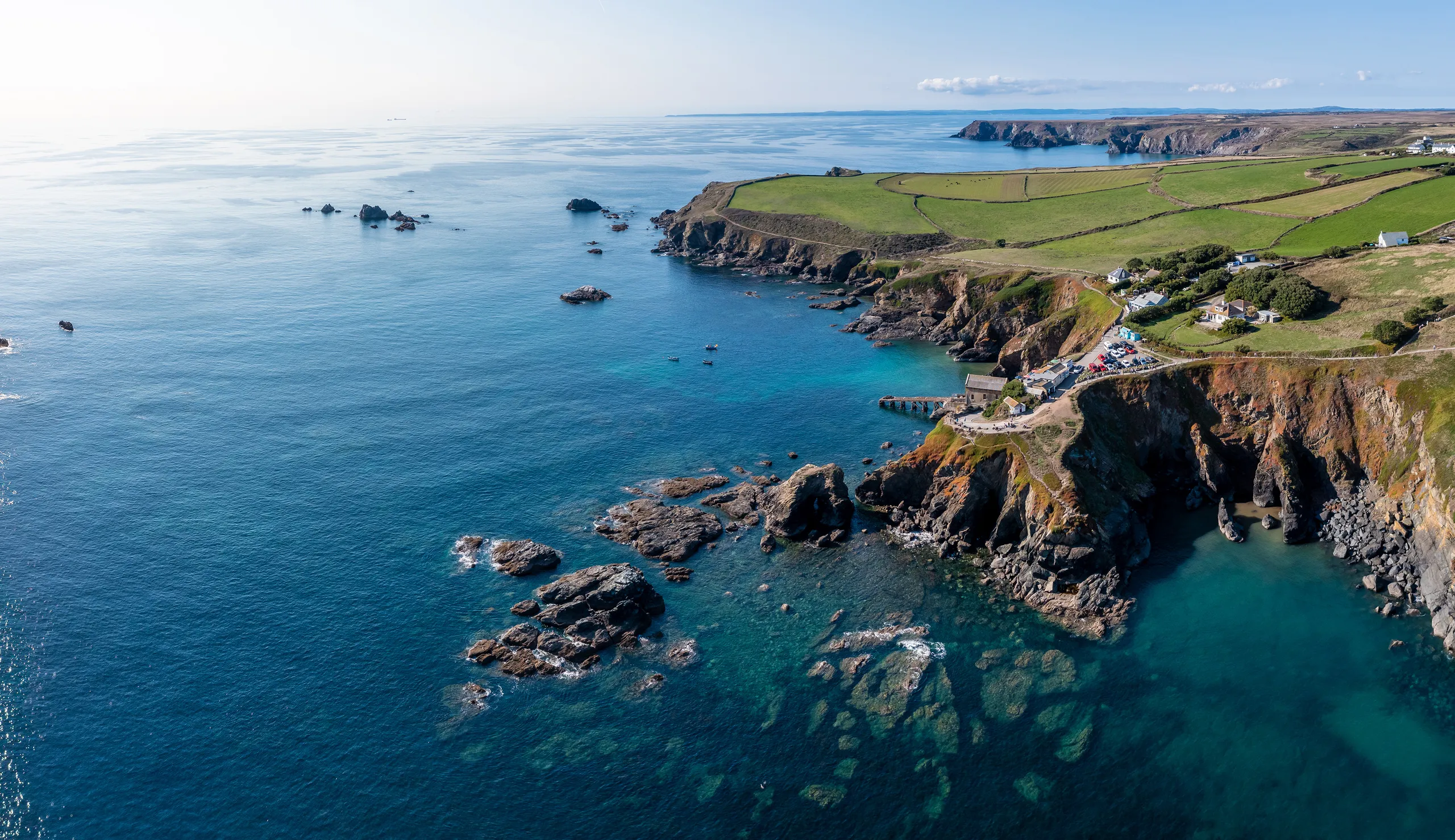 Aerial view of a rugged coastal bay with turquoise water, rocky cliffs, scattered houses and green fields stretching along the shoreline.