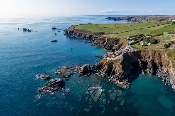 Aerial view of a rugged coastal bay with turquoise water, rocky cliffs, scattered houses and green fields stretching along the shoreline.