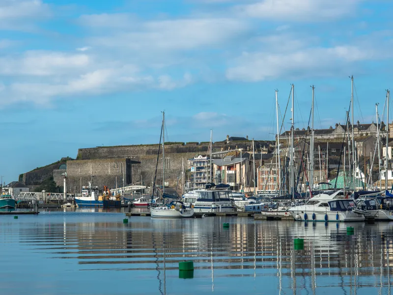 Marina with sailboats and waterfront buildings beside a stone fort under a blue sky