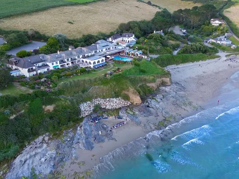 Aerial view of a coastal hotel overlooking a sandy cove with turquoise sea, cliffs and surrounding countryside.