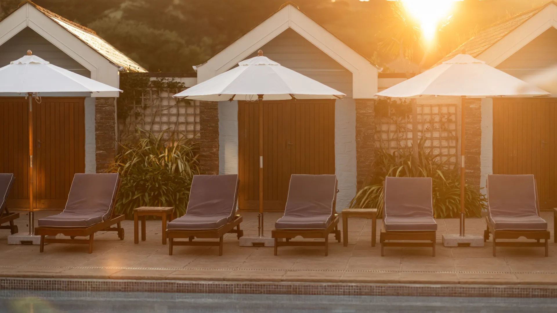 Sun loungers and white parasols beside an outdoor pool at sunset, with poolside cabins in the background.