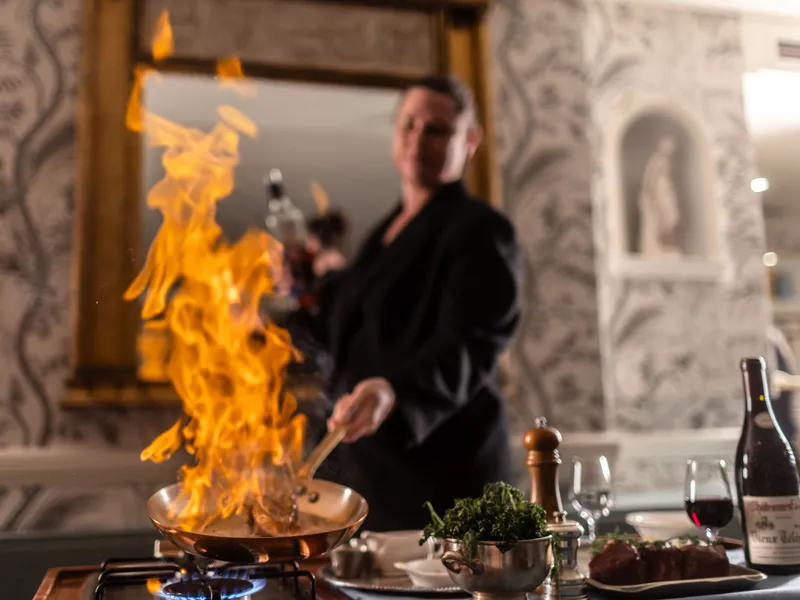 Chef preparing a flambéed dish in a copper pan at a restaurant table, with flames rising high and wine nearby