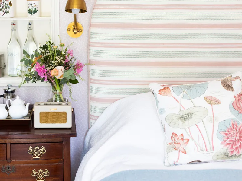 Cosy bedroom corner with a striped upholstered headboard, floral cushion and wooden bedside chest with tea set, flowers and brass wall light