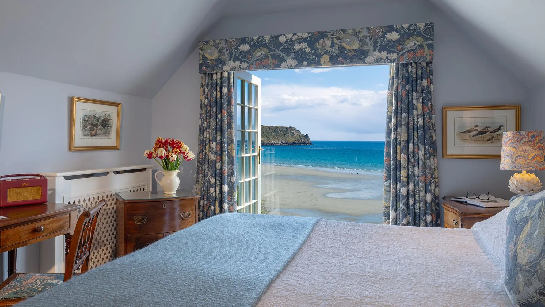 Elegant coastal bedroom with floral furnishings and an open window overlooking a sandy beach and turquoise sea