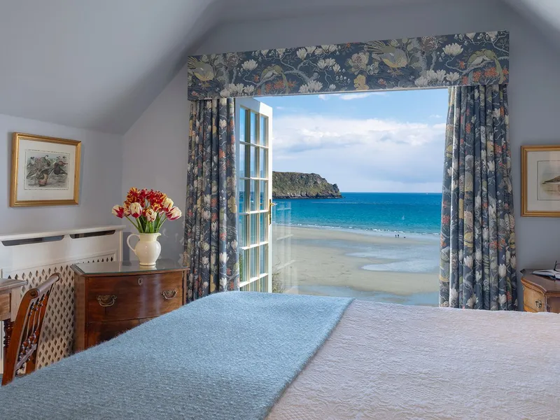 Elegant coastal bedroom with floral furnishings and an open window overlooking a sandy beach and turquoise sea