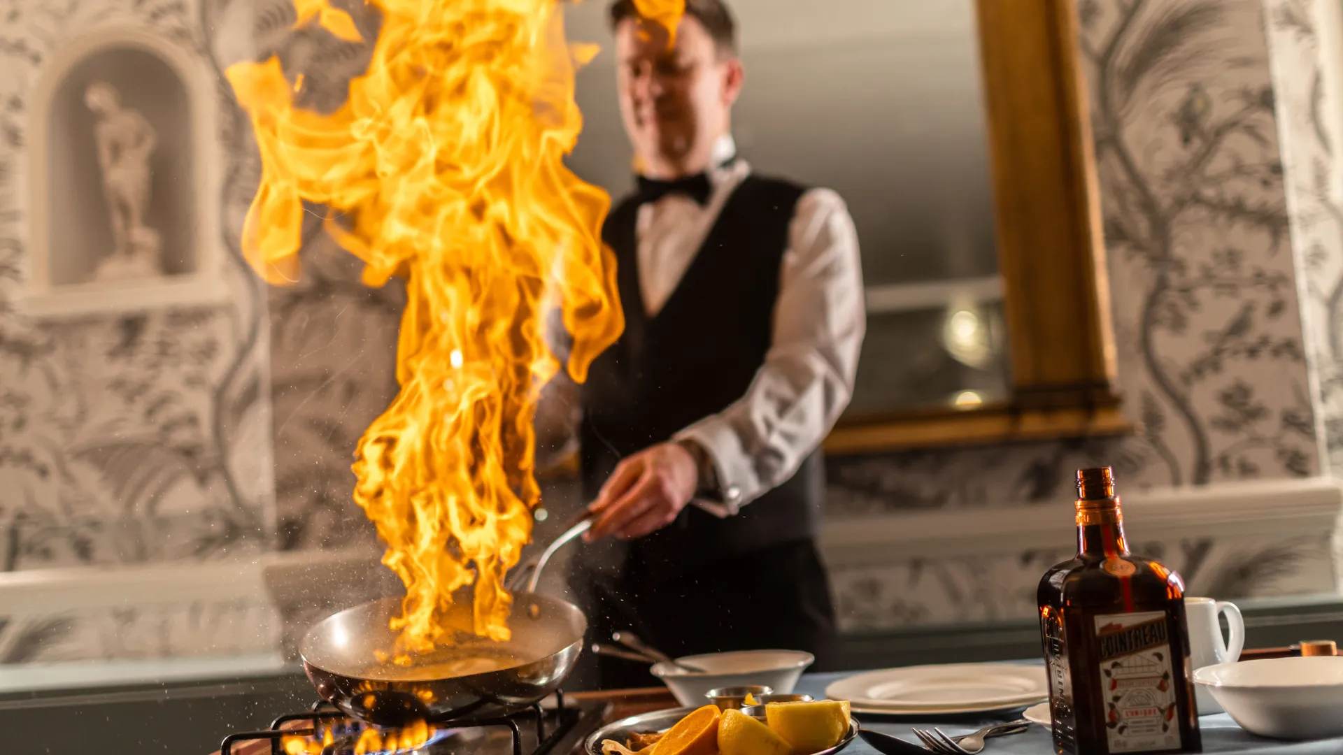 Waiter preparing a flambé dish with tall flames rising from a frying pan beside oranges and liqueur.