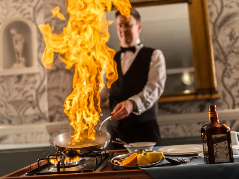 Waiter preparing a flambé dish with tall flames rising from a frying pan beside oranges and liqueur.