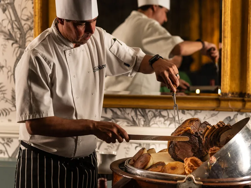 Chef in a tall white toque carving a large roast at a dining room trolley