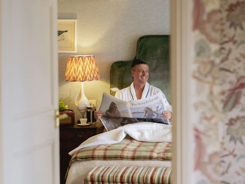 Man in a white dressing gown sitting up in bed reading a newspaper in a cosy bedroom