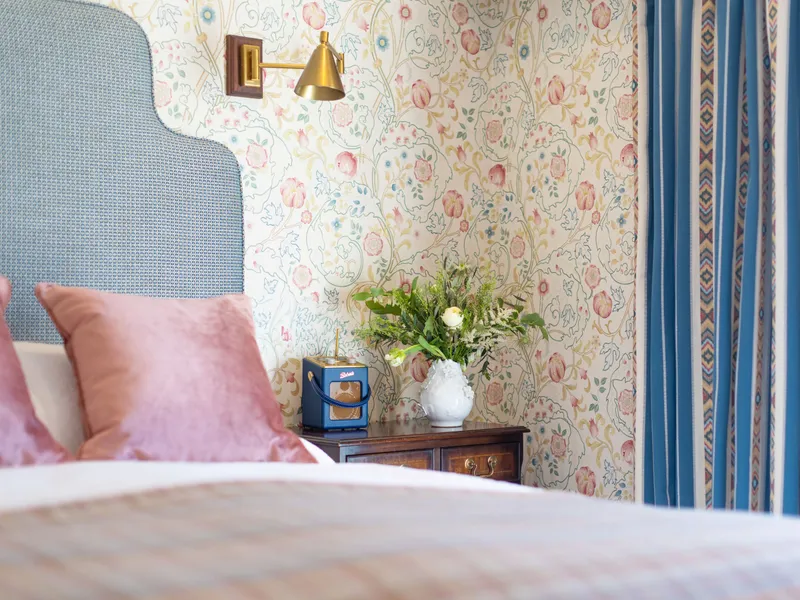 Elegant bedroom corner with a blue upholstered headboard, pink velvet cushions, floral wallpaper and blue striped curtains