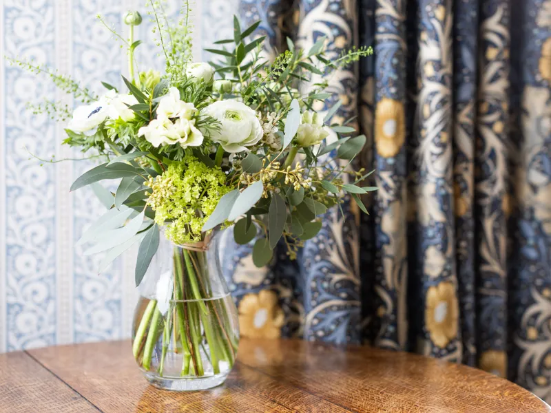Fresh white and green floral arrangement in a clear glass vase on a wooden table, set against patterned blue curtains.